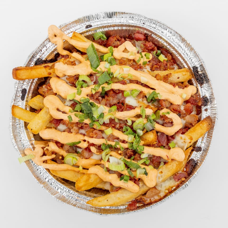 Loaded fries in a foil container, topped with bacon, melted cheese, orange sauce, and green onions.