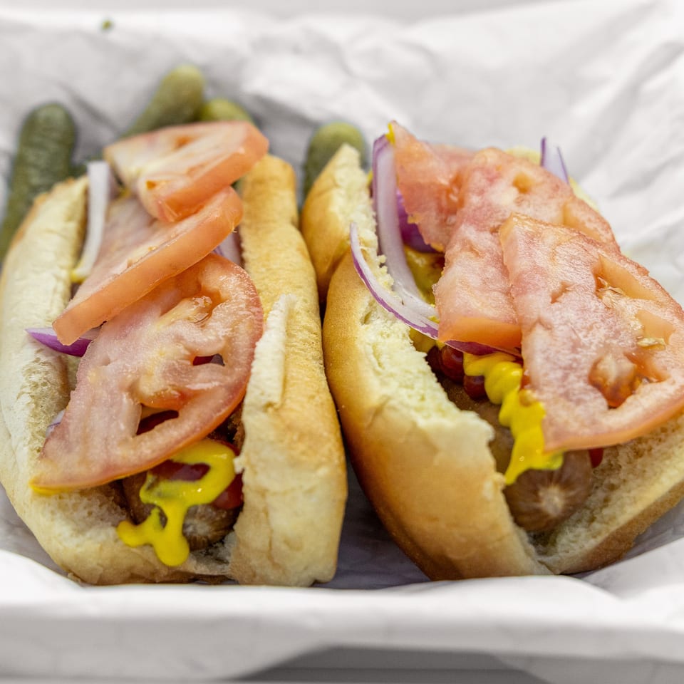 Two hot dogs in buns with sliced tomatoes, red onions, mustard, and pickles in a white paper tray.